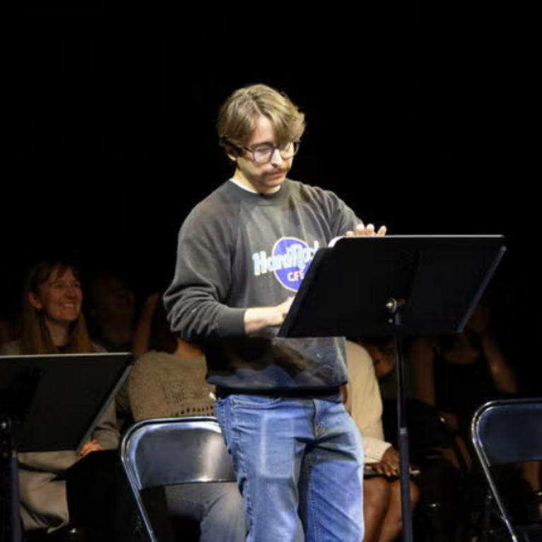 three students performing in front of music stands and other students in the background watching