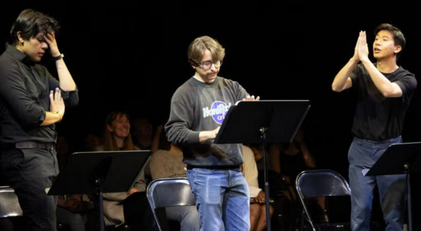 three students performing in front of music stands and other students in the background watching